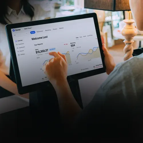 Person using a touchscreen tablet showing a dashboard with sales report of $15,368.37 and returning customers at 504.